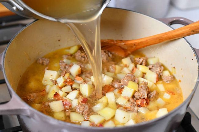 pouring chicken stock into soup pot with diced potatoes and Italian sausage