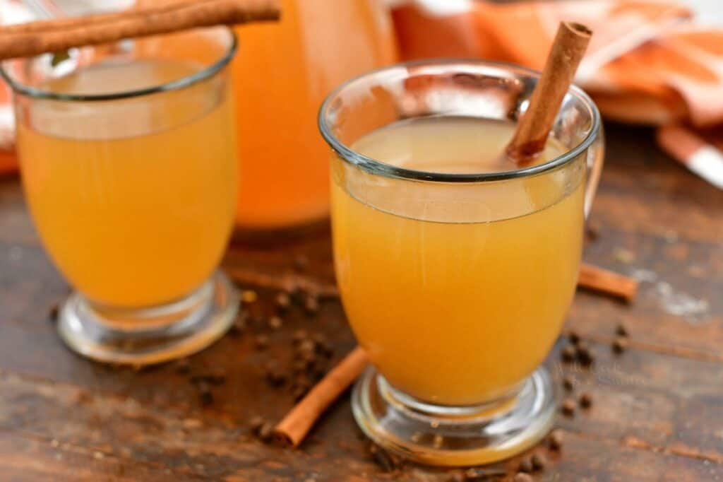 side view: two clear glass mugs of apple cider with cinnamon sticks