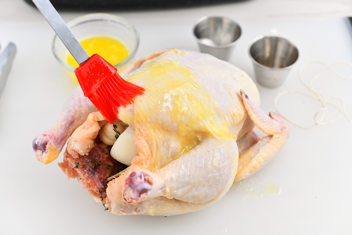 using red silicone basting brush to apply melted butter onto uncooked whole chicken