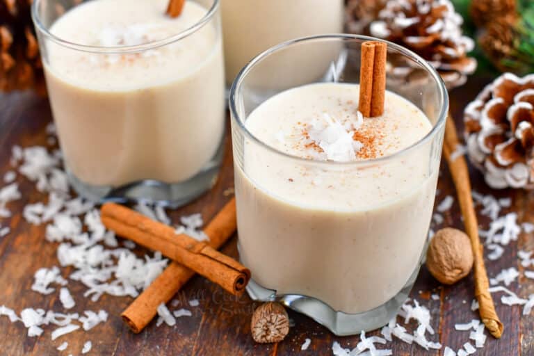 Three glasses of creamy eggnog garnished with cinnamon sticks and a sprinkle of coconut flakes sit on a wooden surface. Cinnamon sticks and whole nutmeg surround the glasses, with pine cones in the background, creating a festive atmosphere.
