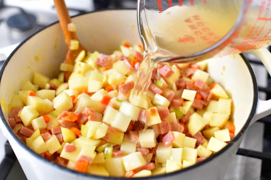 adding chicken stock to a pot of vegetables, potatoes and ham for soup recipe