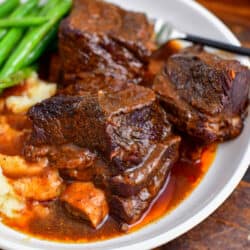 plate of three soft and tender short ribs recipe with sauce mashed potatoes and green beans.