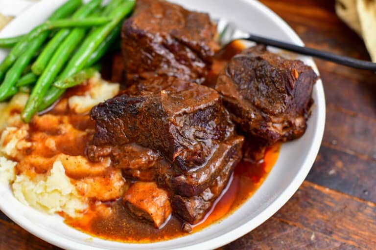 plate of three soft and tender short ribs recipe with sauce mashed potatoes and green beans.