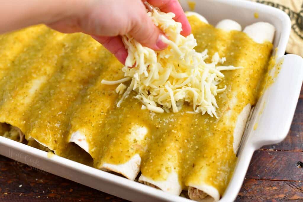 hand sprinkling shredded cheese over uncooked green chicken enchiladas