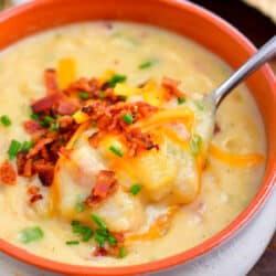 creamy soup topped with bacon, shredded cheese and chives in a white and orange bowl with a spoon