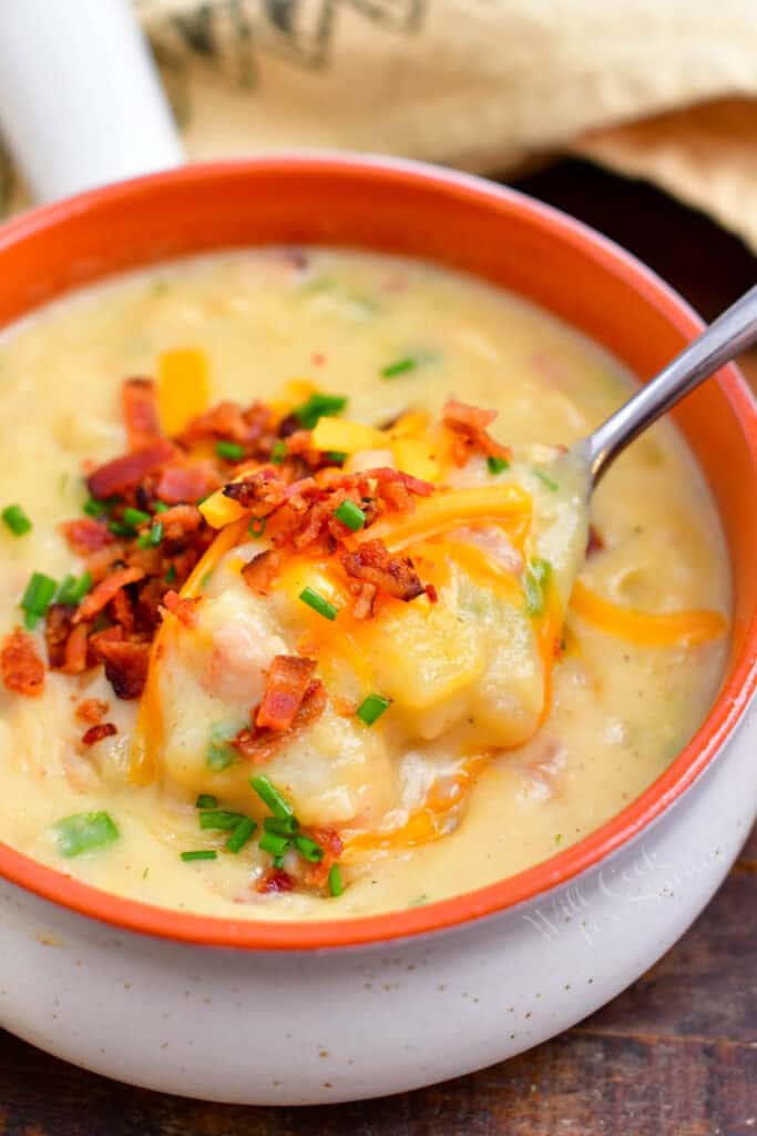 creamy soup topped with bacon, shredded cheese and chives in a white and orange bowl with a spoon