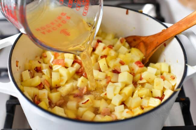 broth being poured from a glass measuring cup into a pot of potatoes and chopped bacon