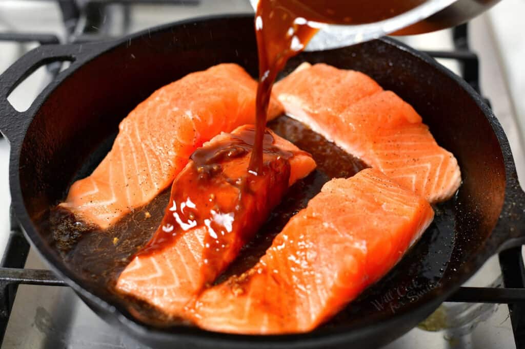 teriyaki sauce being poured over salmon fillets in a cast iron skillet