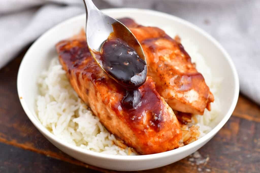 spooning sauce onto teriyaki salmon in white dish on bed of jasmine rice