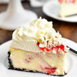 slice of cheesecake with frosting, raspberry sauce, and white chocolate curls on top with cake stand in the background