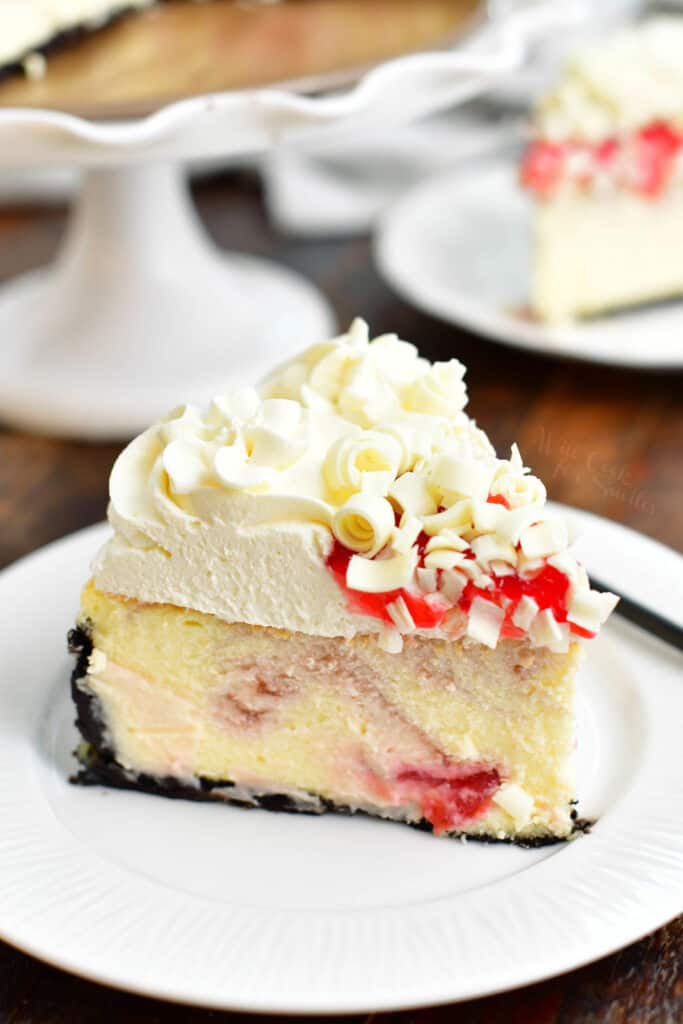 slice of cheesecake with frosting, raspberry sauce, and white chocolate curls on top with cake stand in the background