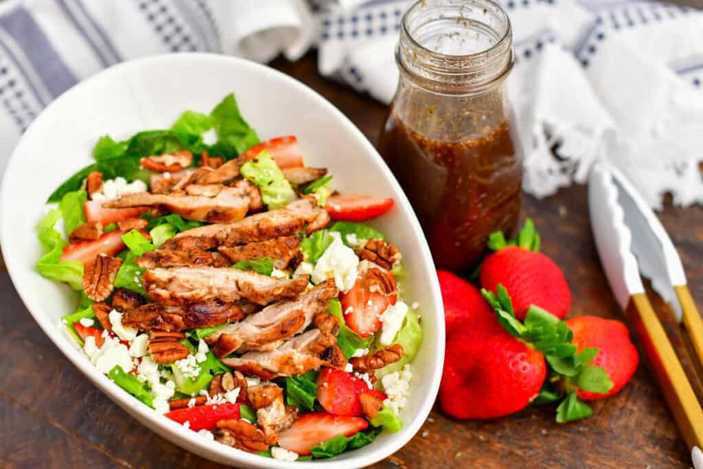 bowl of salad with sliced chicken surrounded by patterned towels, jar of dressing, strawberries, and a pair of tongs