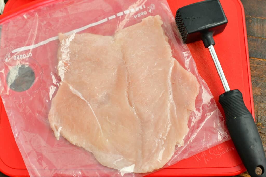 flattened chicken breast on red cutting board next to a meat mallet