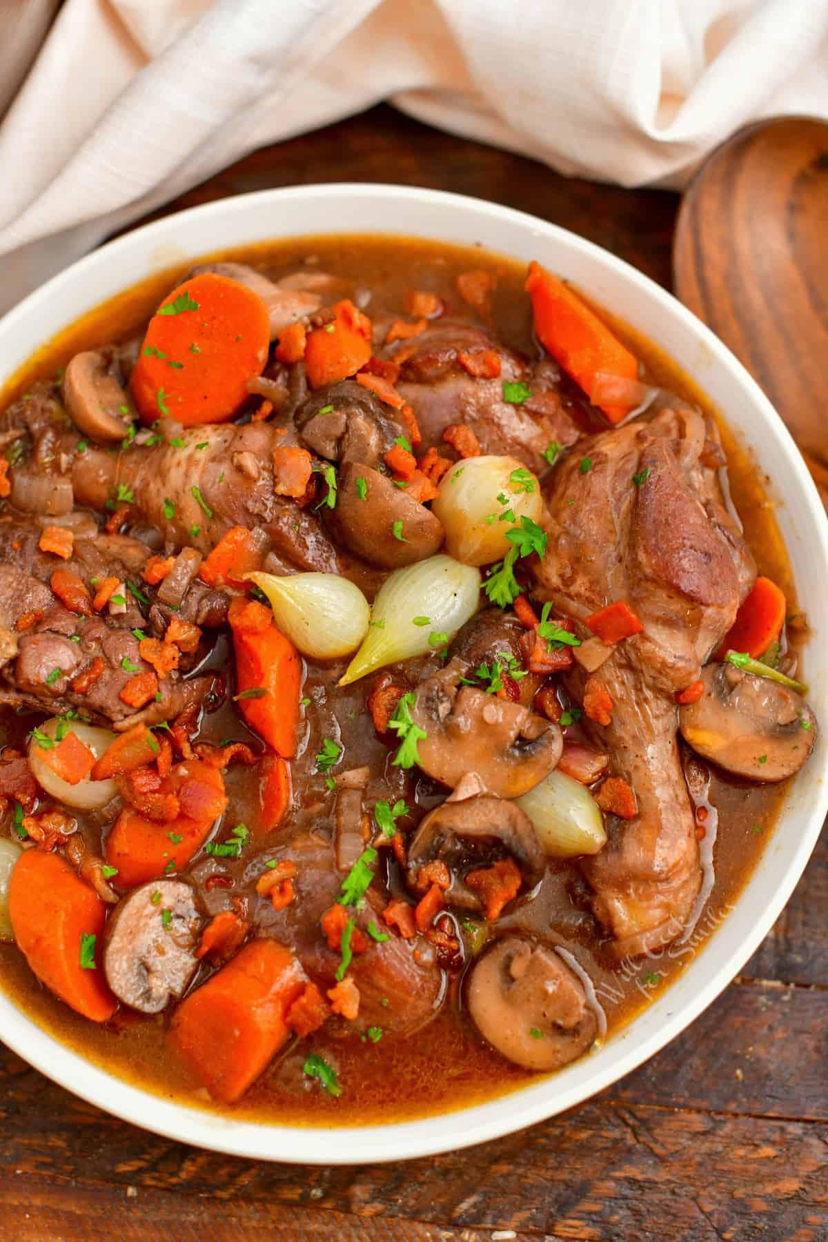 overhead: white bowl of coq au vin on a wooden surface surrounded by a white cloth and wooden spoon