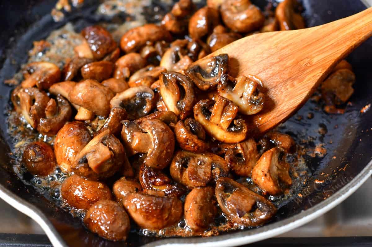 stirring with a wooden spoon buttery cut mushrooms sauteed in a skillet.