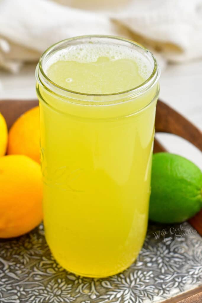 tall glass jar of sweet and sour mix surrounded by lemons and limes