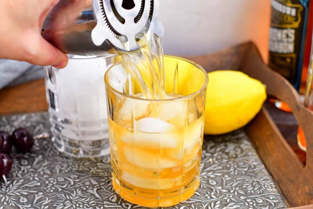 An amaretto sour has been shaken, and is now being poured through a cocktail strainer into a glass half filled with ice. 