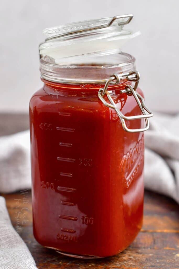 A jar with a half open lid is filled with dark red barbecue sauce.