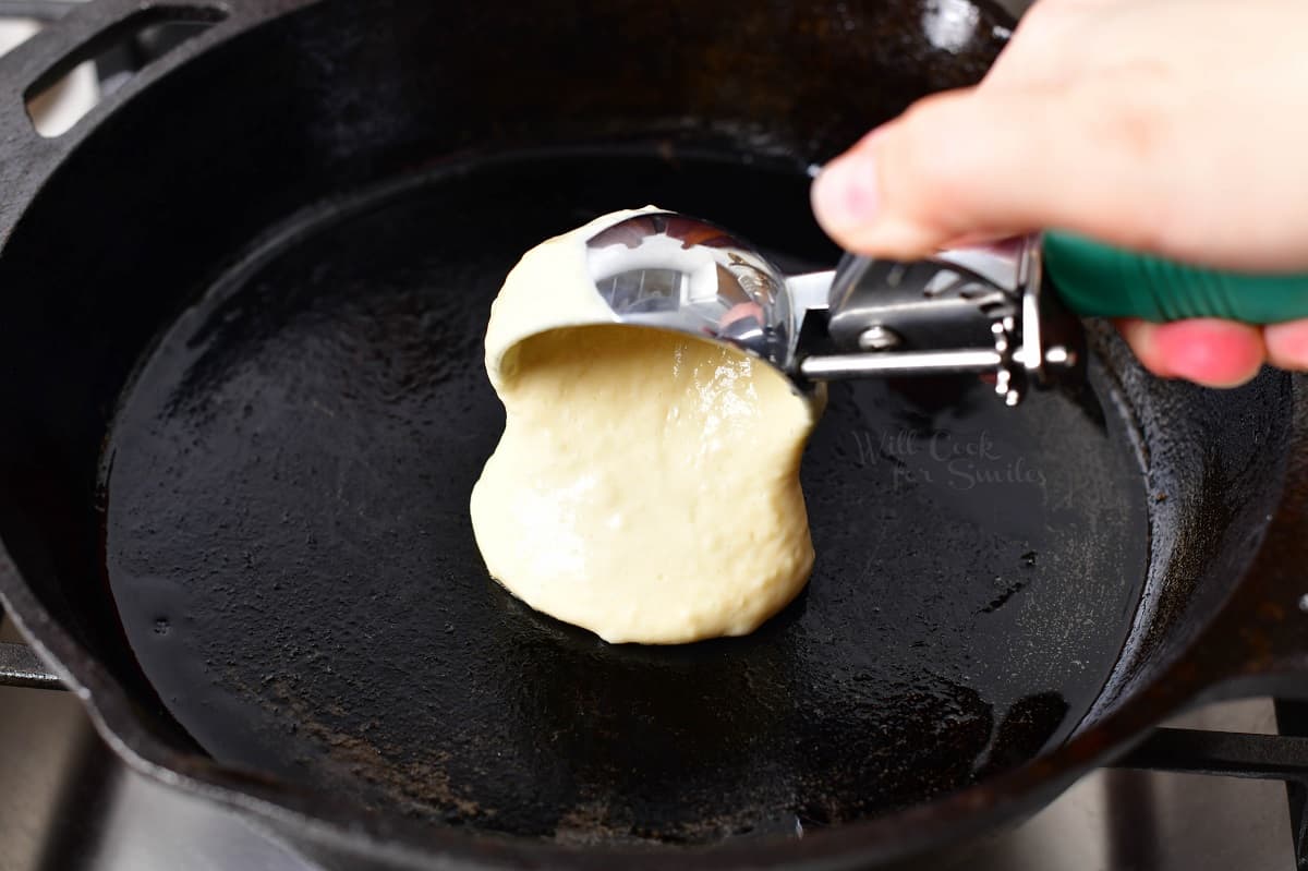 An ice cream scoop is placing pancake batter onto a hot skillet. 