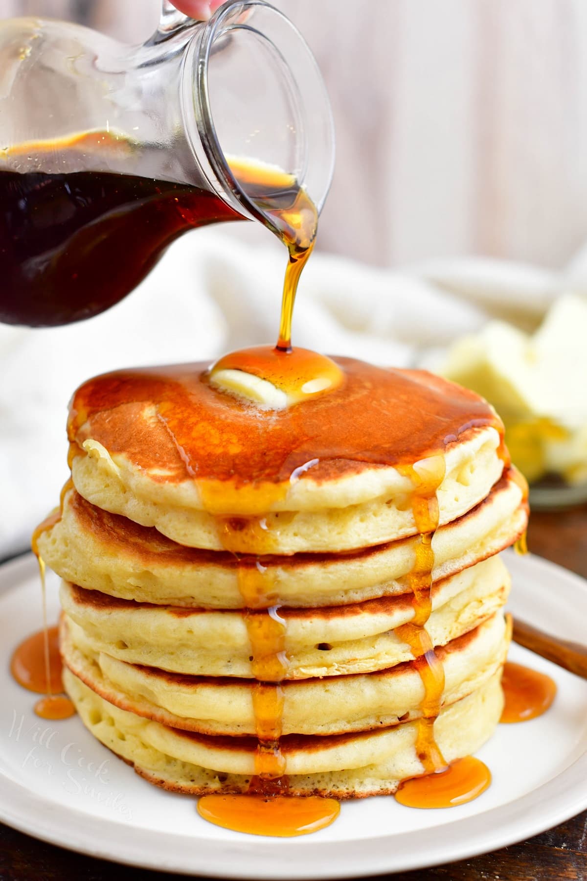 Maple syrup is being poured over a stack of buttermilk pancakes.