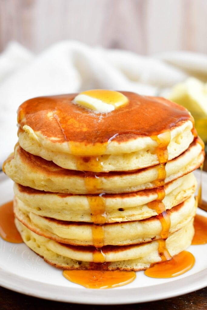 A stack of buttermilk pancakes is presented on a white plate with butter and syrup.