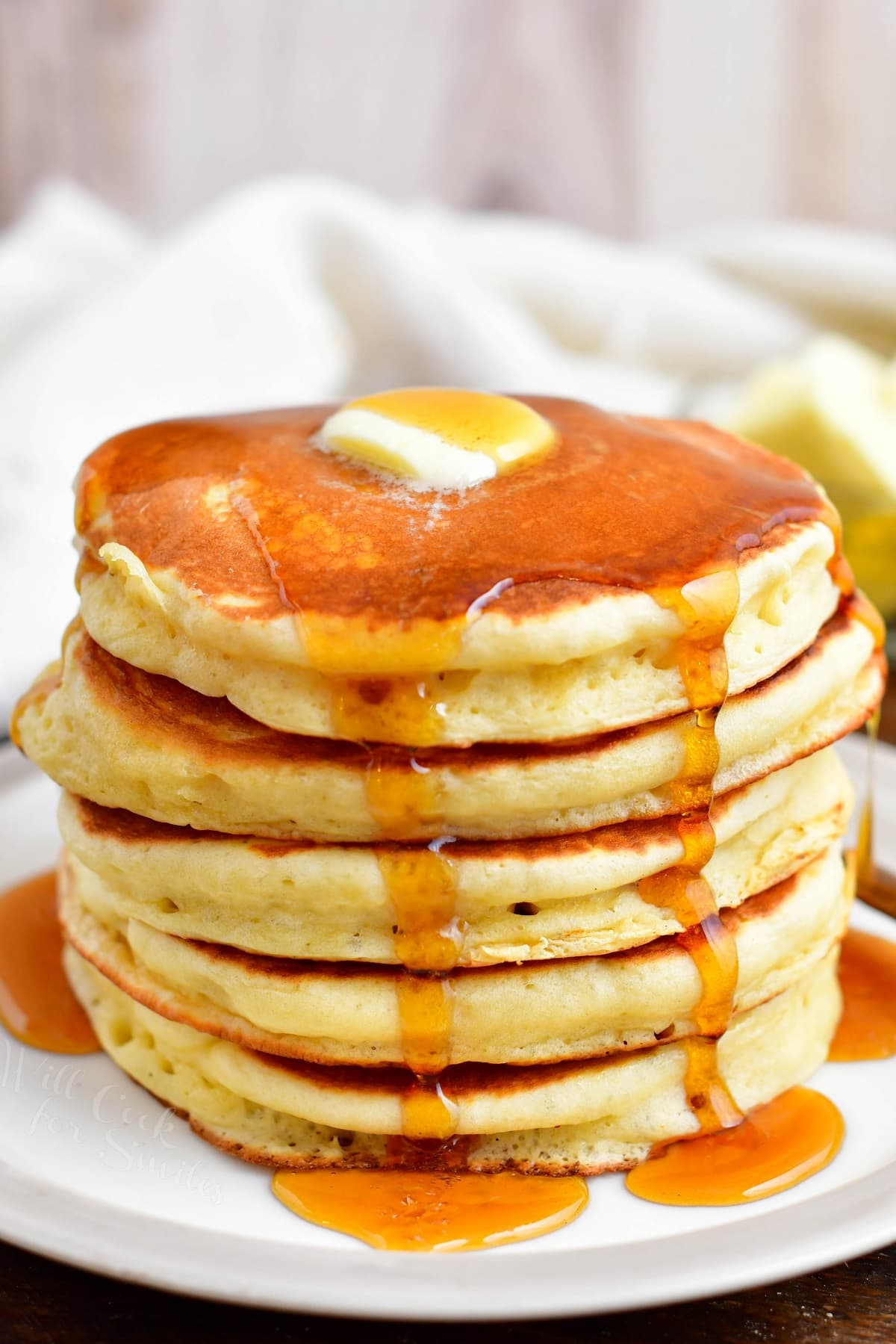 A stack of buttermilk pancakes is presented on a white plate with butter and syrup.
