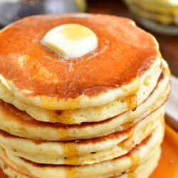 A stack of buttermilk pancakes is topped with a square of butter and drizzled with maple syrup.