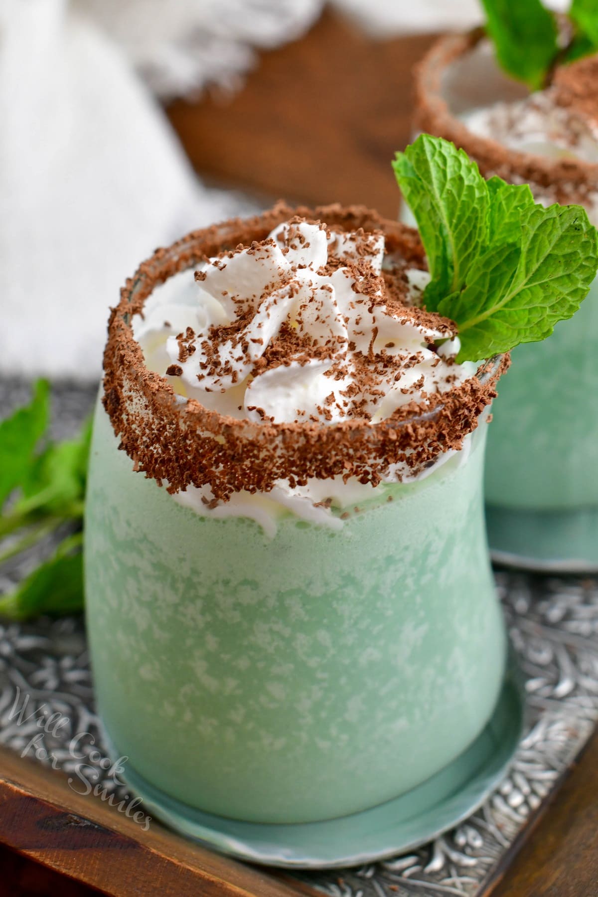 A green cocktail is presented on a metal surface with chocolate and whipped topping.