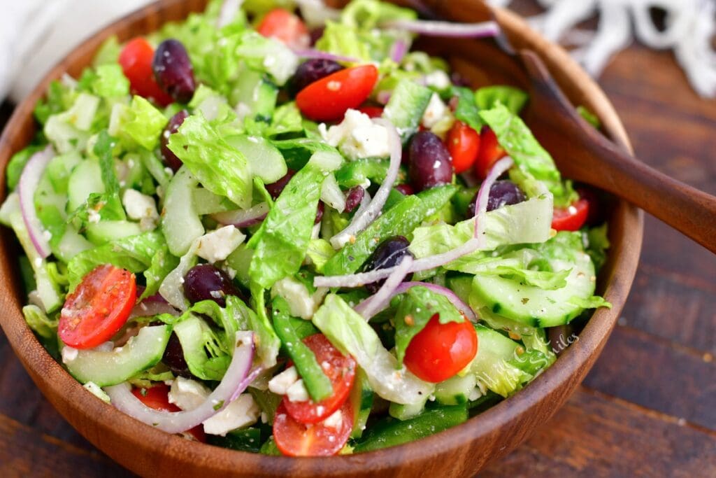 A freshly made greek salad is in a wooden bowl with a large wooden serving spoon.