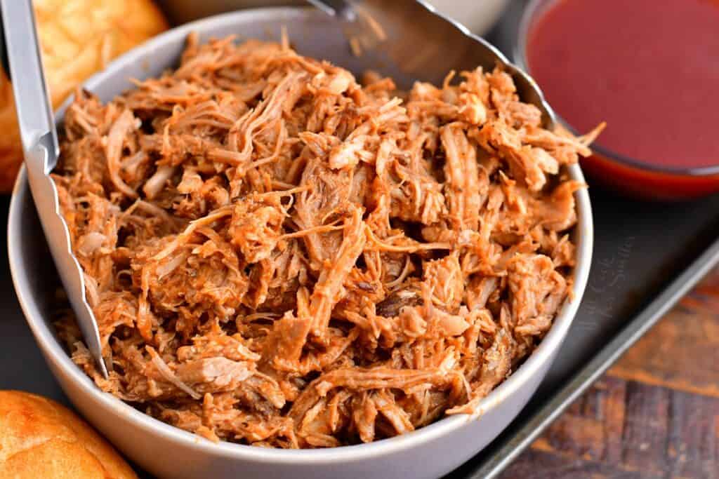 Tongs are placed in a bowl full of pulled pork.