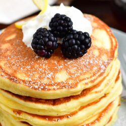 A stack of ricotta pancakes is topped with whipped cream and blackberries.