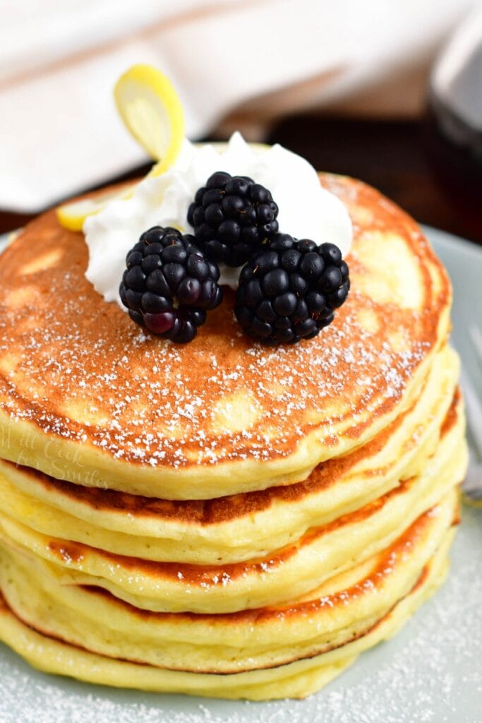 A stack of ricotta pancakes is topped with whipped cream and blackberries.