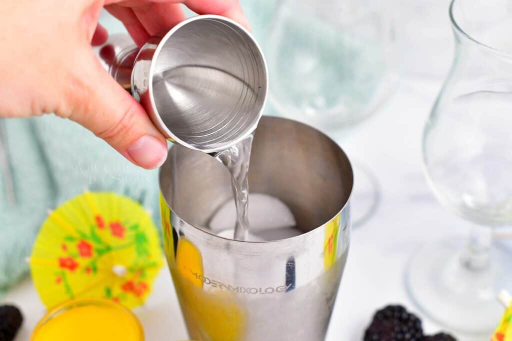 A shot of white rum is being poured into a silver cocktail shaker.