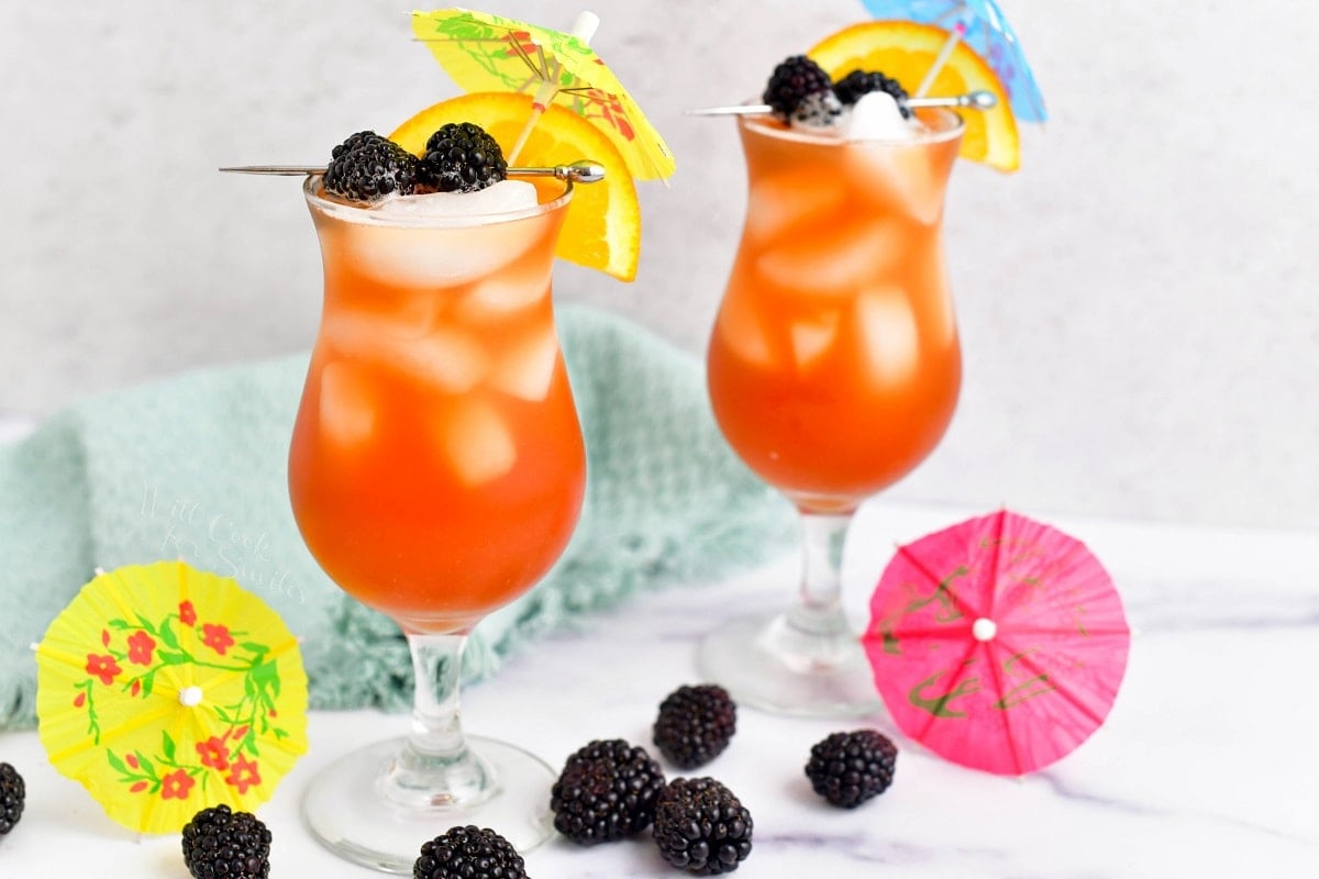 Two hurricane glasses are filled with orange rum runner and garnished with fruit and umbrellas.