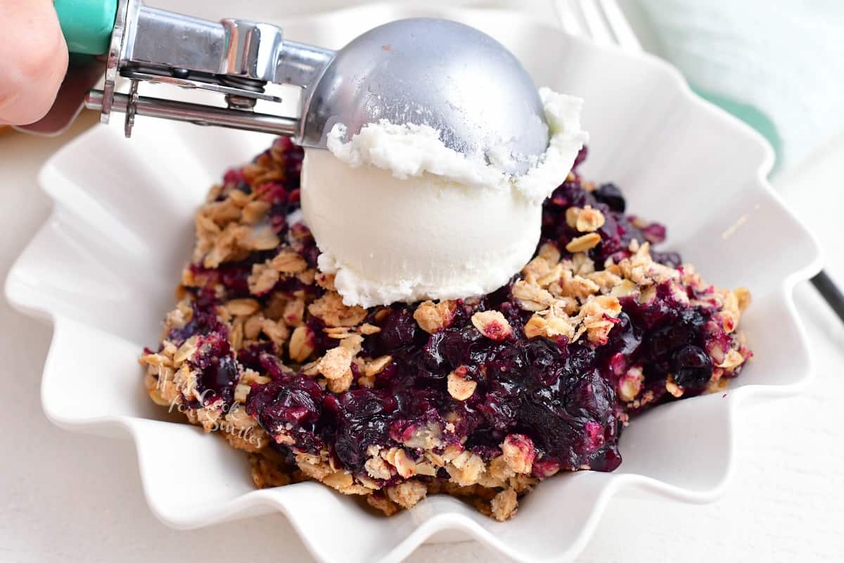 An ice cream scooper is placing a scoop of vanilla ice cream onto a serving of blueberry crisp.