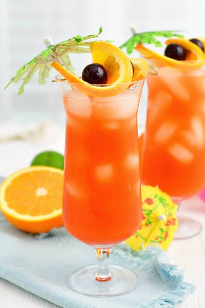 A tall glass of rum runner is garnished with fruits and small palm trees.