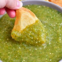 A triangular tortilla chip is being dipped into a large bowl of salsa verde.