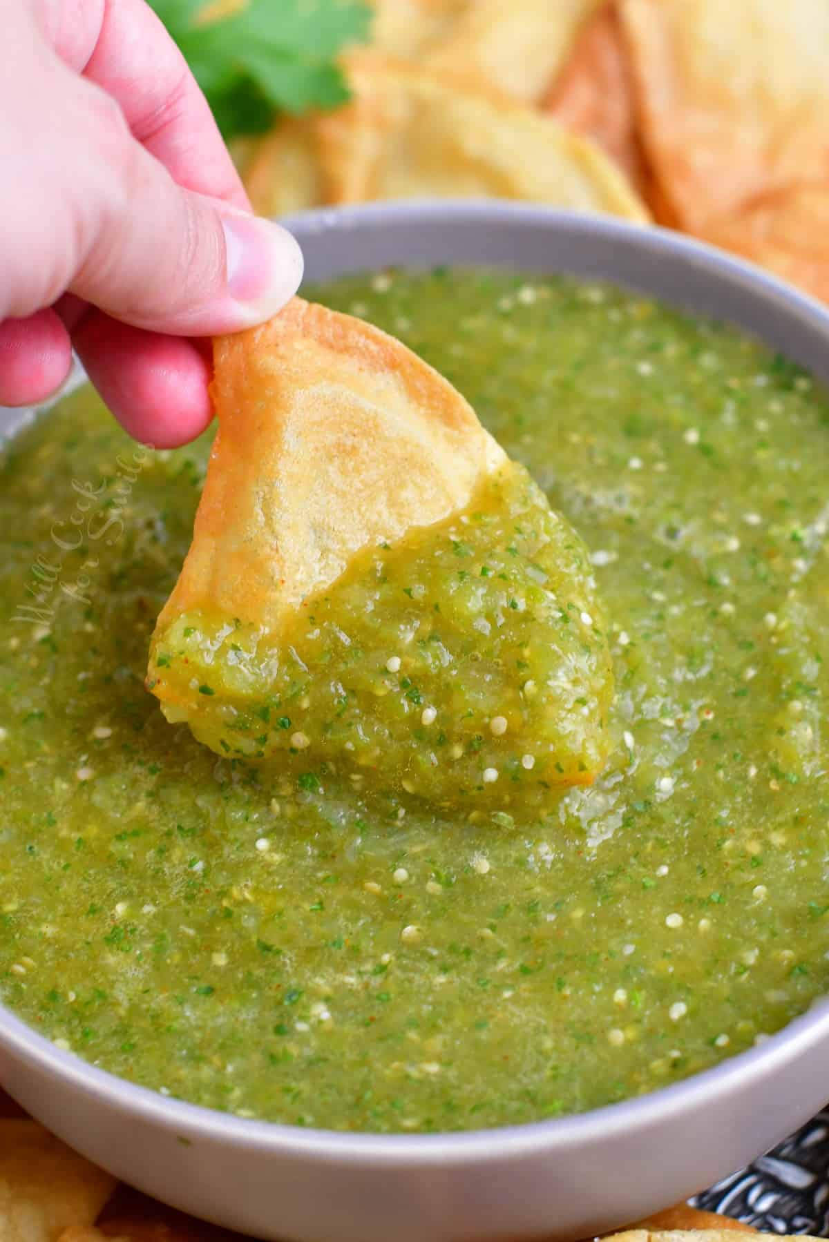A triangular tortilla chip is being dipped into a large bowl of salsa verde.