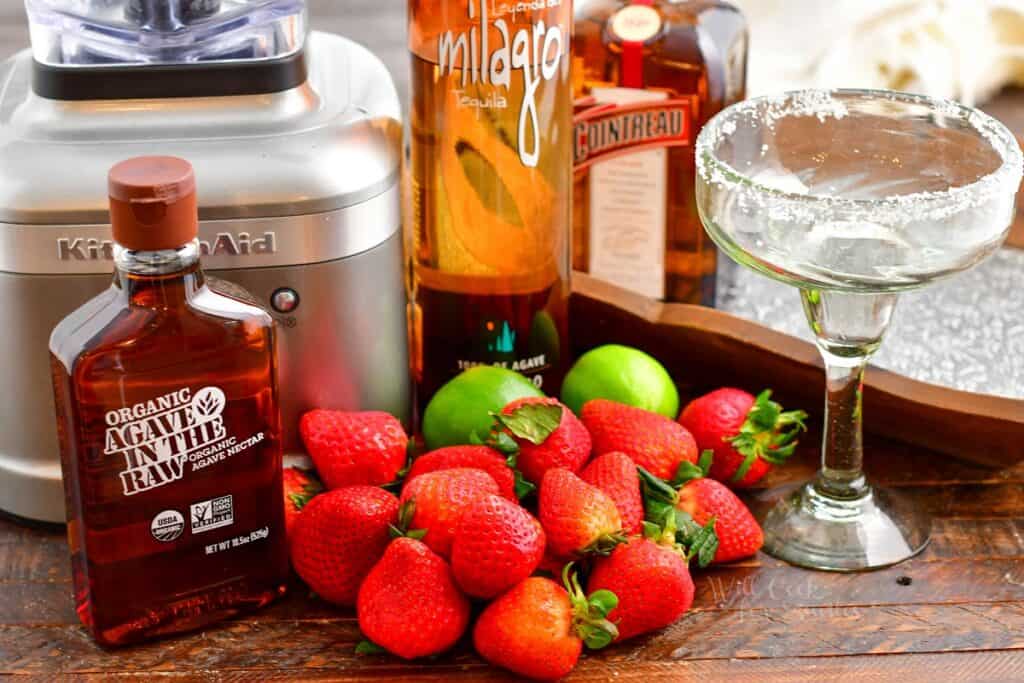 The ingredients for strawberry margaritas are laid out on a wooden surface. 