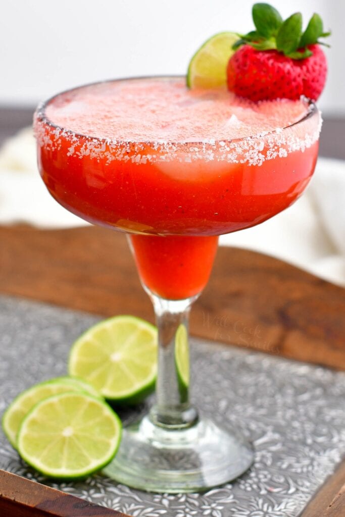 Slices of lime are next to a tall glass of strawberry margarita. 