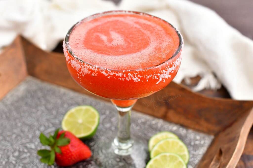 Limes and a strawberry surround a glass of strawberry margarita. 