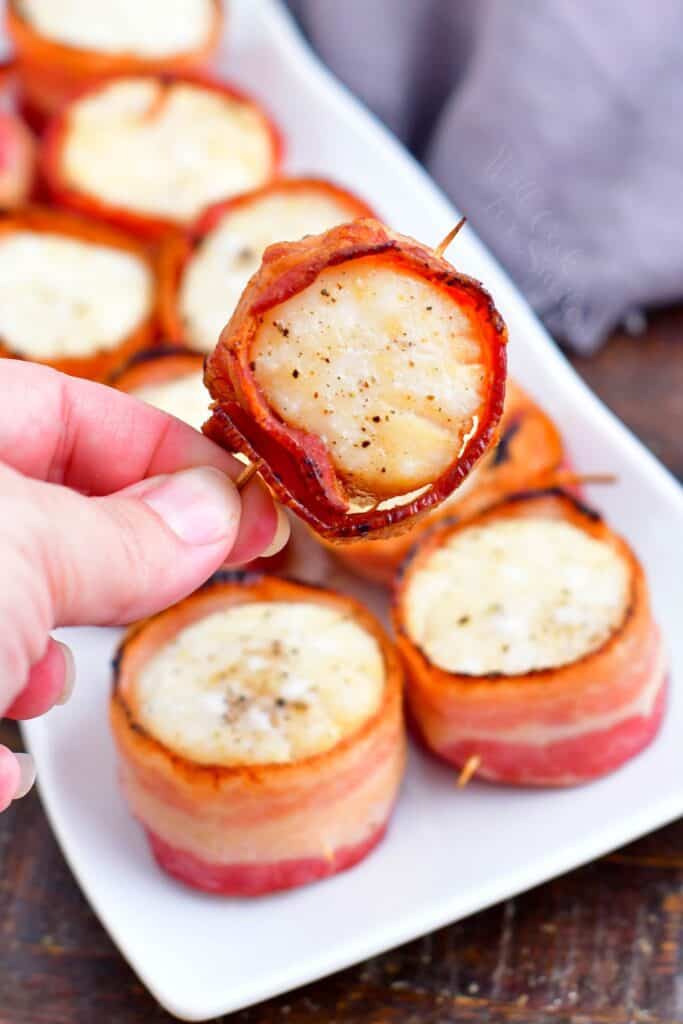 A bacon wrapped scallop is lifted from a white plate.