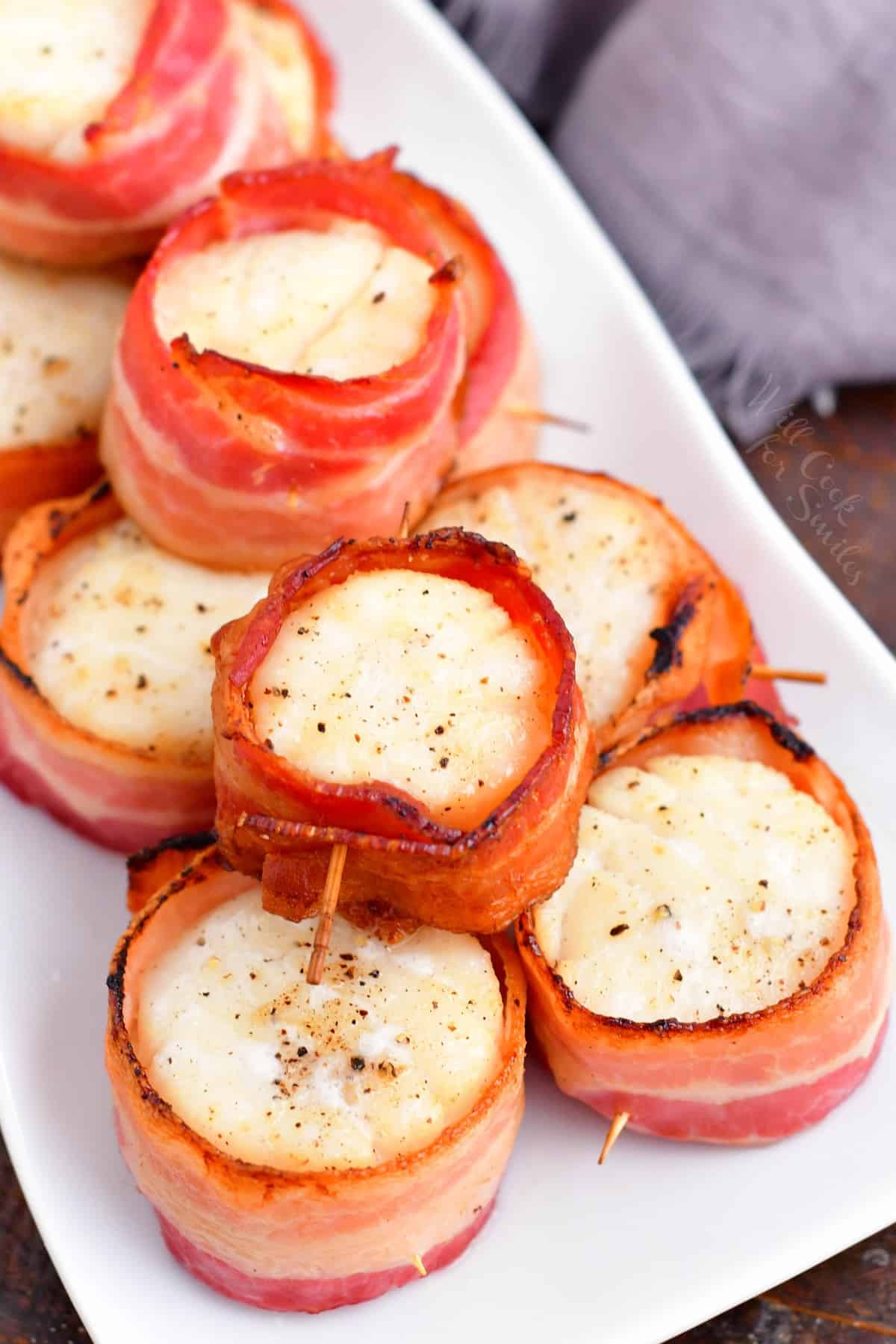 Scallops wrapped in bacon are piled on a rectangular white plate.