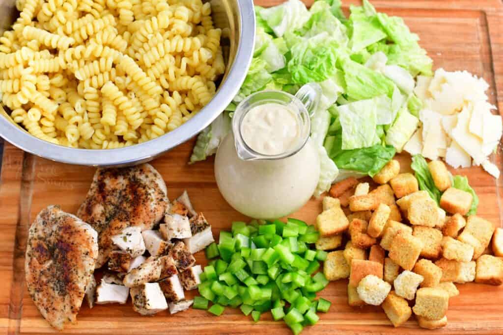 The ingredients for chicken caesar pasta salad are placed on a wooden surface. 