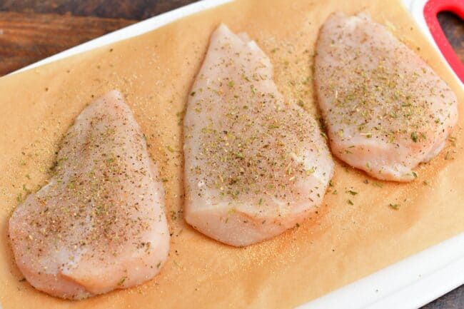 The raw chicken breasts are seasoned on a sheet of parchment paper. 