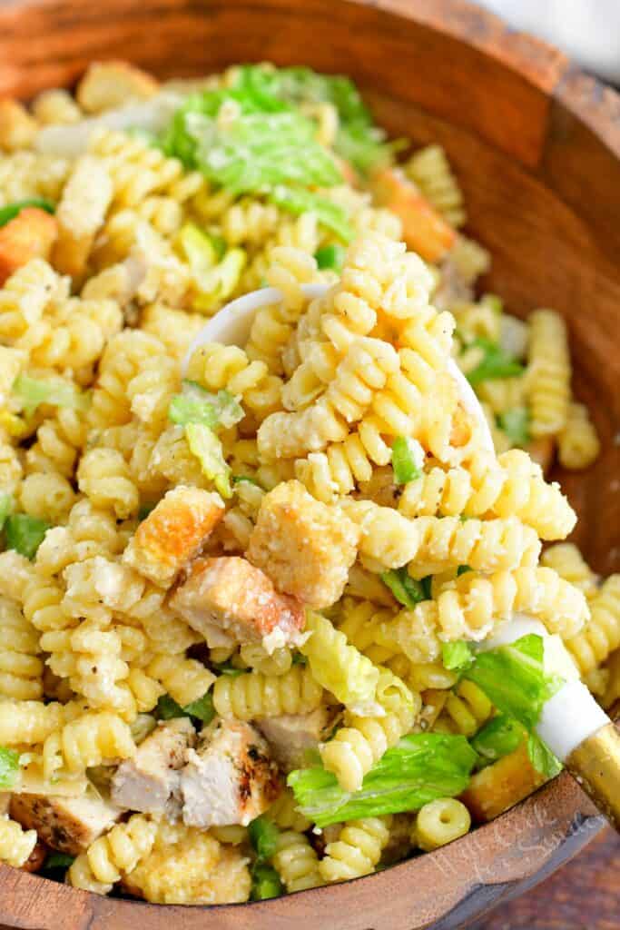 A serving spoon is lifting a portion of pasta salad from a large bowl. 