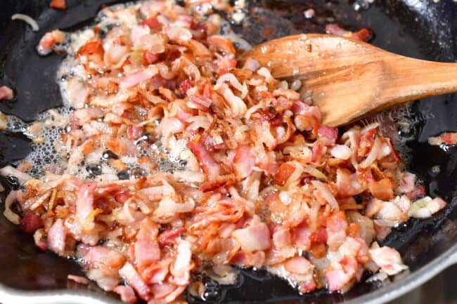 Bacon is being cooked in a black skillet with a wooden spoon. 
