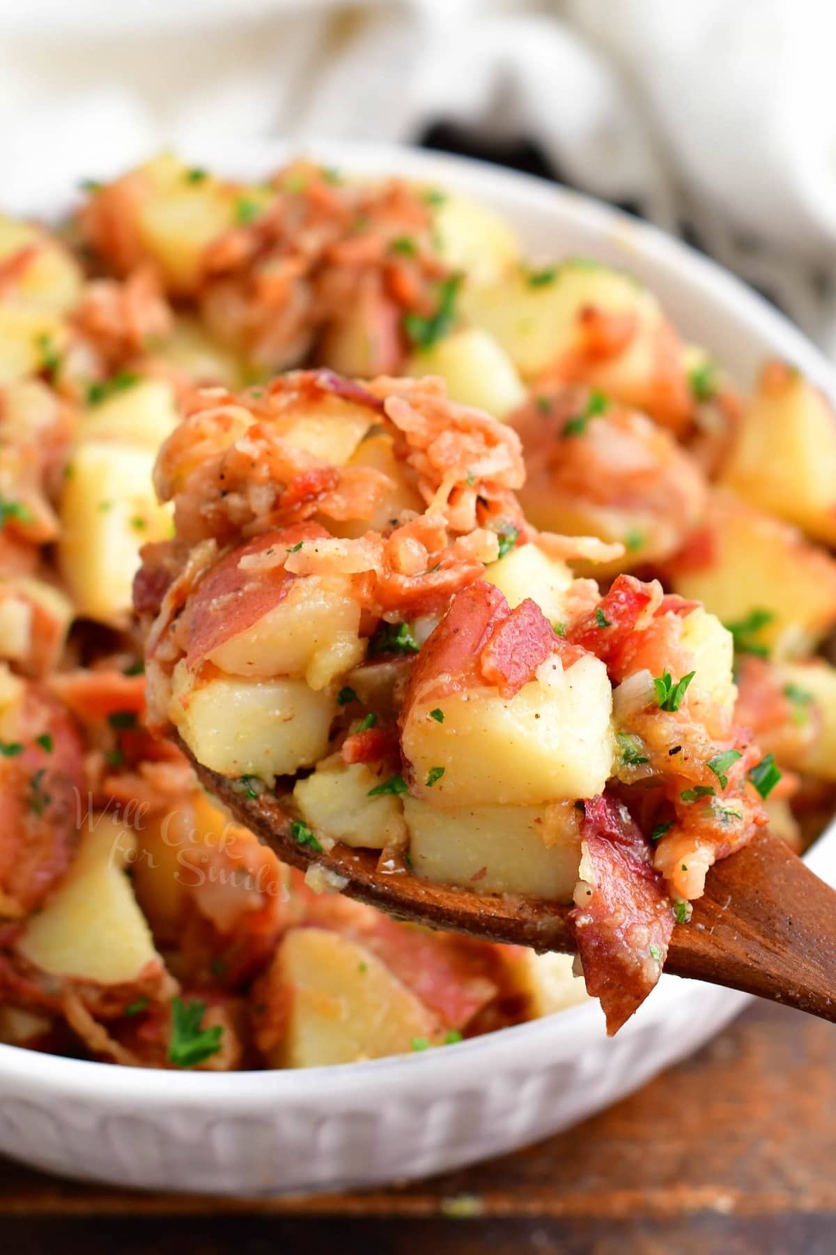 A wooden spoon is lifting a portion of potato salad from the bowl. 