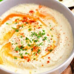 A white bowl filled with hummus is ready to be eaten.