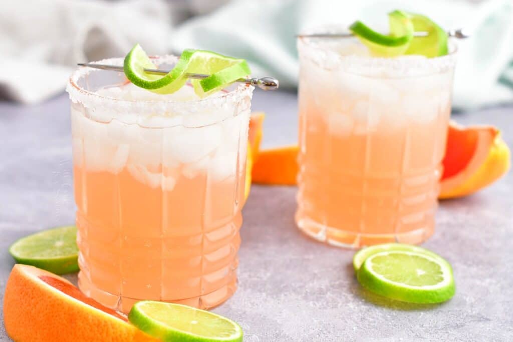 Two palomas are placed next to a few lime wedges on a counter.